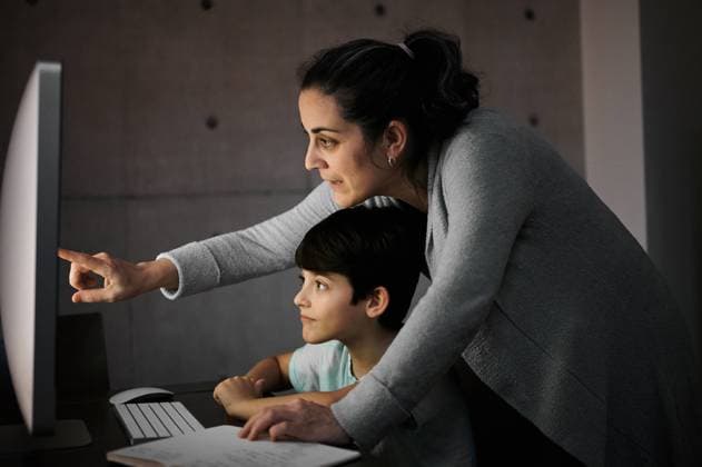IMAGO Creative: Gesellschaft, Integration Side view of young woman explaining study task to positive son sitting at table with computer and textbook during online lesson at home, Model released Copyright: xJuanxGarciaxRisquezx MR