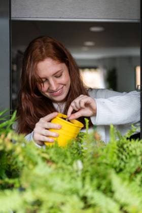 Lovely young female smiling and planting seed in small yellow pot while standing on balcony of stylish apartment Copyright: xKikexArnaizx Model Released