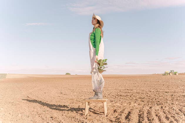 IMAGO Creative: Klimawandel - Steigende Temperaturen Young woman standing on stool on dry field, carrying plant in a net model released Symbolfoto ERRF02315