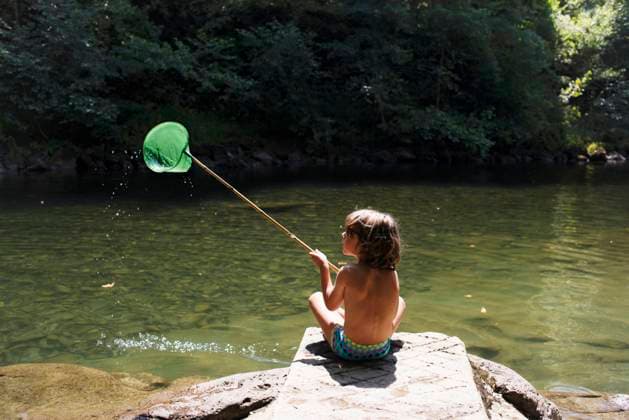 Back view of shirtless little boy sitting on stone and catching fish with skoop net while spending summer holidays at riverside Copyright: xManuelxRuizxAlbax Model Released