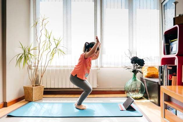 Side view of positive little girl standing in yoga pose on mat near window at home while practicing yoga and watching video tutorials on tablet Copyright: xManuelxRuizxAlbax Model Released