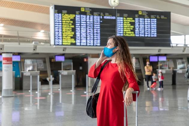 IMAGO Creative: Welttourismustag am 27. September Serious female traveler in mask standing with suitcase on background of departure board and talking on the phone while waiting for flight Copyright: xPabloxGarciax Model Released