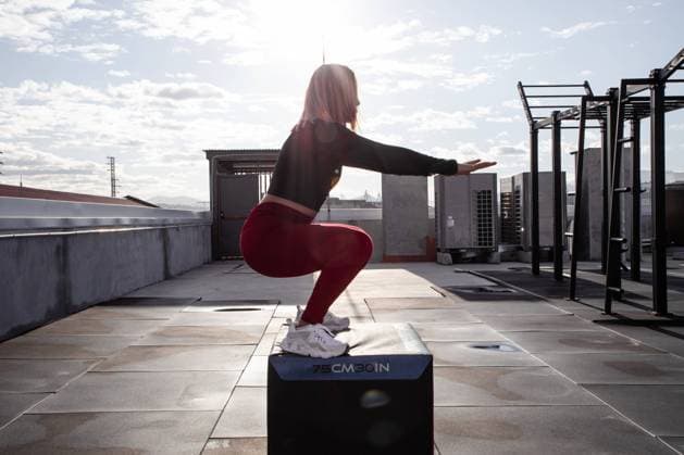 IMAGO Creative: Sport in der Stadt Side view of concentrated woman in sportswear jumping on box and squatting while training on rooftop with modern sport equipment in city Copyright: xPabloxGarciax Model Released
