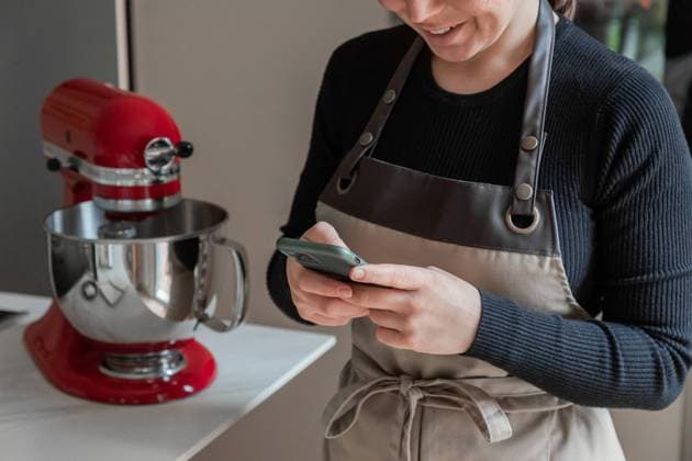 IMAGO Creative: Haushaltsger?te - K?chenmaschine Crop positive smiling housewife in apron using mobile phone and reading recipe while standing next to table with red stand mixer in home kitchen Copyright: xPabloxGarciax Model Released
