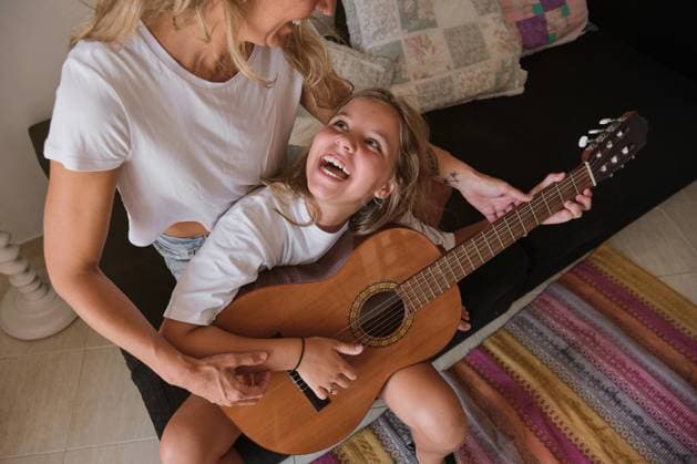 IMAGO Creative: Bildung, Musikerziehung Blonde woman teaching a girl to play the guitar while looking at each other laughing sitting on a couch in a house, Model released Copyright: xPhilippexDegrootex MR