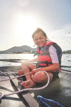 Vertical photo of a blond kid with happy expression in a life jacket sitting in a boat holding the rudder and a rope to drive a boat with the shore far away in the background, Model released Copyright: xPhilippexDegrootex MR
