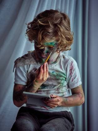 IMAGO Creative: Ruhe im Chaos Cute funny boy painted in bright paints with mirror sitting near curtains and drawing on face Copyright: xPhilippexDegrootex Model Released