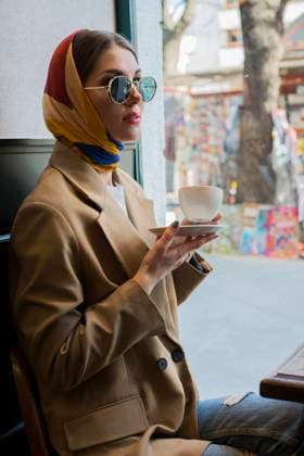 Side view of trendy young female in eyeglasses and shawl on head enjoying coffee while sitting with crossed legs in cafeteria near window in afternoon Copyright: xSebastianxCandiollix Model Released