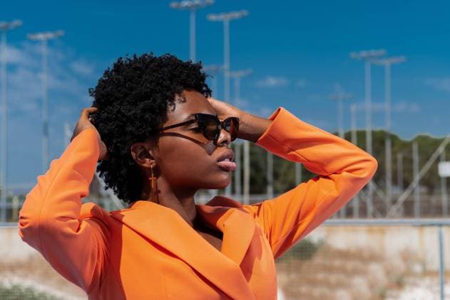 IMAGO Creative: Clickbait, Modetrends From below of young contemplative black female in trendy orange clothes and sunglasses looking away with raised arms under blue cloudy sky, Model released Copyright: xSergioxMenax MR