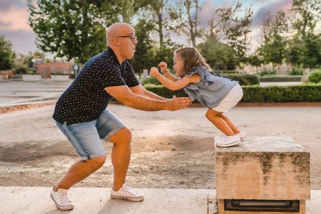 IMAGO Creative: Sommer, Sommertage im Stadtpark Side view of father standing in park and ready for catching playful daughter jumping from stone parapet while entertaining together in city, Model released Copyright: xSixAndxSix MR