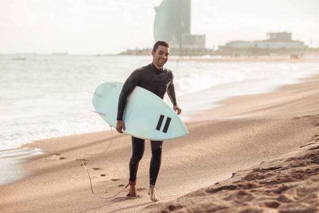 IMAGO Creative, Jahreszeit Sommer, Sommer in der Stadt Happy adult man in wetsuit carrying surfboard walking on sandy beach in sunlight, Barcelona Copyright: xAitorxLamadridx Model Released