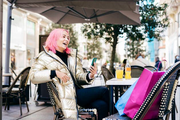 IMAGO Creative: Urban Diversity Pink hair woman using mobile phone while sitting with shopping bags at sidewalk cafe model released Symbolfoto MARF00024