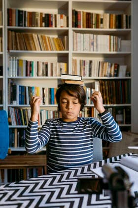 IMAGO Creative: NGO, Ausbildung für Kinder Boy balancing books on head while vlogging video camera in living room model released Symbolfoto property released VABF04063
