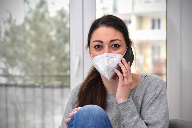 Eine junge Frau sitzt in ihrer Wohnung und arbeitet im Homeoffice wegen der Corona Pandemie.Sie traegt eine FFP2 Maske und telefoniert mit einem Smartphone. MODEL RELEASED!