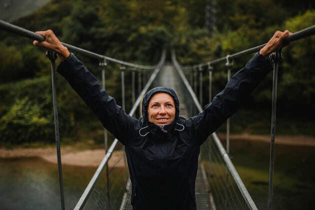 IMAGO Creative: Ende des Sommers Creative Highlights Symbolbilder Smiling woman wearing raincoat with arms raised standing on suspension bridge over Sella river in forest model released Symbolfoto DMGF00421