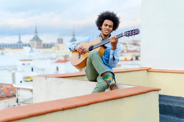 IMAGO Creative: Bildung, Musikerziehung Tag der Musik am 21. Juni Young man with curly hair playing acoustic guitar while sitting on terrace model released Symbolfoto PGF00320