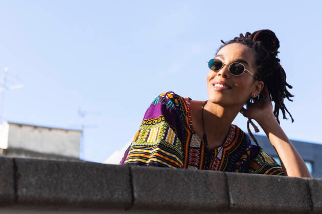 IMAGO Creative: Clickbait, Modetrends Woman with dreadlocks wearing sunglasses looking away while leaning on retaining wall against sky model released Symbolfoto PNAF00436
