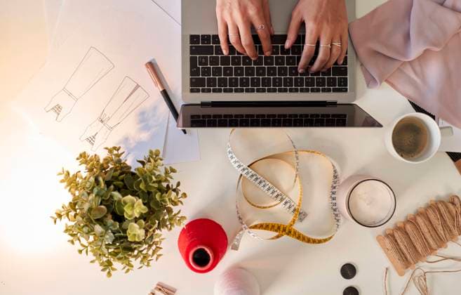 Fashion designer using laptop by fabric swatch and coffee at desk in atelier model released Symbolfoto property released VEGF03439