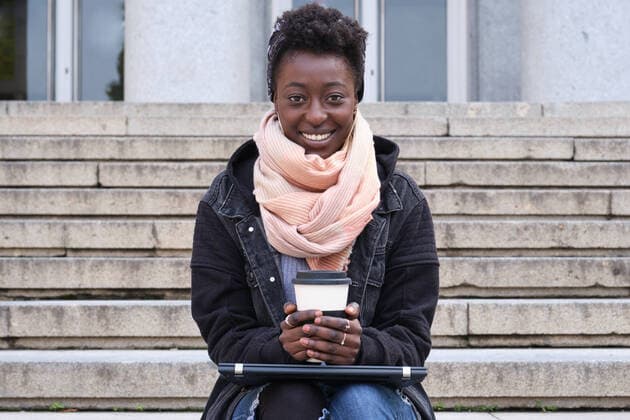 Beautiful university female african student smiling looking at camera, holding a cup of coffee, sitting on stairs outside on campus. College life concept. Madrid, Community of Madrid, Spain CR_CRVI210111-599706-01 ,model released, Symbolfoto