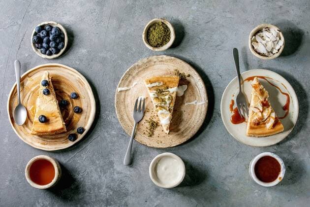 Creative Highlights Symbolbilder Pieces of cheesecake with different toppings on ceramic plates and variety of sauces in ceramic bowls in row over grey texture background. Flat lay, space ÂNatashaBreen REDA&CO 1028_29_NBR8491