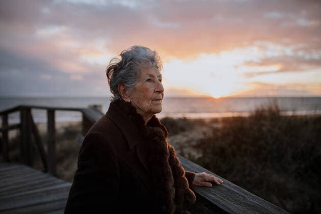 Side view of elderly female traveler in casual clothes standing on wooden pier on sandy beach and enjoying seascape at sunset Copyright: xAlbertoxOrtegax Model Released