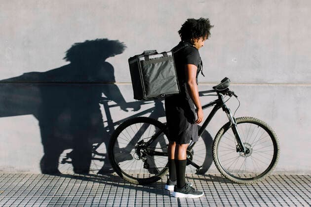 Side view of black delivery rider with box and bike standing on city pavement against wall with shadow while looking down in sunlight Copyright: xLightsyx Model Released