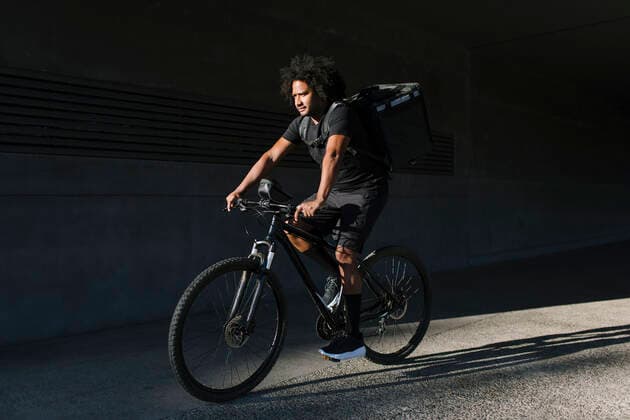 IMAGO Creative, Sommer, Schatten-Pl?tze bei Hitze Side view of ethnic male with delivery box riding bicycle on walkway against wall with shadow in sunlight Copyright: xLightsyx Model Released
