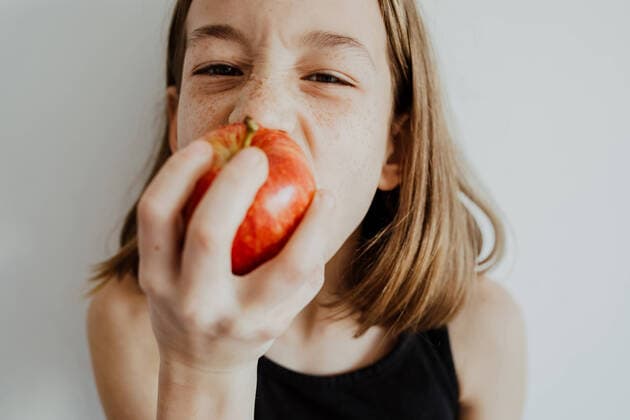 IMAGO Creative: 2021 Favoriten Girl in casual top looking at camera while biting fresh ripe red apple against white background Copyright: xSunsixAlbetsx Model Released
