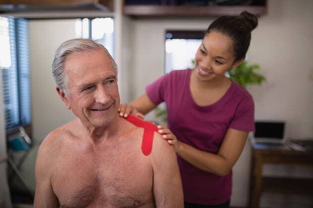 Female therapist applying elastic therapeutic tape on shoulder of smiling shirtless senior male pati Female therapist applying elastic therapeutic tape on shoulder of smiling shirtless senior male patient at hospital ward, model released, , property released, Cape Town, South Africa, 23.04.2017, 1034624.jpg, physiotherapy, physical therapy, medical clinic, healthcare, alternative therapy, care, hospital, examination room, ward, lifestyle, patient, recovery, treatment, therapist, physiotherapist, doctor, support, healing, female doctor, multi-ethnic group, practitioner, specialist, health service, expertise, healthcare worker, millennial, occupation, service occupation, profession, therapy, muscle retraining, touching, examining, bandage, s