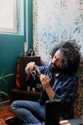 Hipster man holding camera while sitting cross legged against wall at home model released Symbolfoto property released MRRF00847