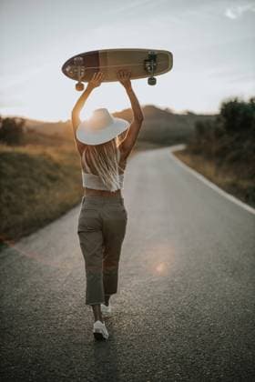 IMAGO Creative: Sommer, Abendspaziergang Back view young female in casual wear and summer hat holding cruiser skateboard and looking away while standing on empty asphalt road in rural area at sunset, Model released Copyright: xDavidxFernandezx MR