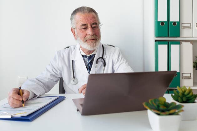 Senior male doctor in uniform writing memos on paper during online health consultation on portable computer in hospital Copyright: xSergioxVictorxVegax Model Released