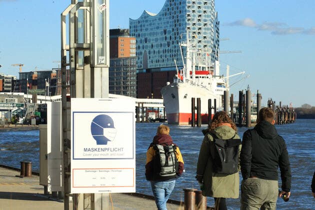 Bei den St. Pauli Landungsbrücken an der Elbe ist ein Schild angebracht, das darauf hinweist, dass in diesem Bereich am Wochenende und an Feiertagen von 10 bis 18 Uhr Maskenpflicht besteht. St. Pauli Hamburg
