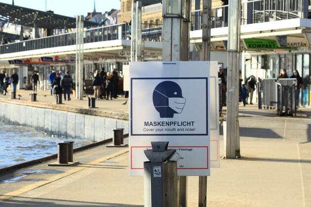Bei den St. Pauli Landungsbrücken an der Elbe ist ein Schild angebracht, das darauf hinweist, dass in diesem Bereich am Wochenende und an Feiertagen von 10 bis 18 Uhr Maskenpflicht besteht. St. Pauli Hamburg