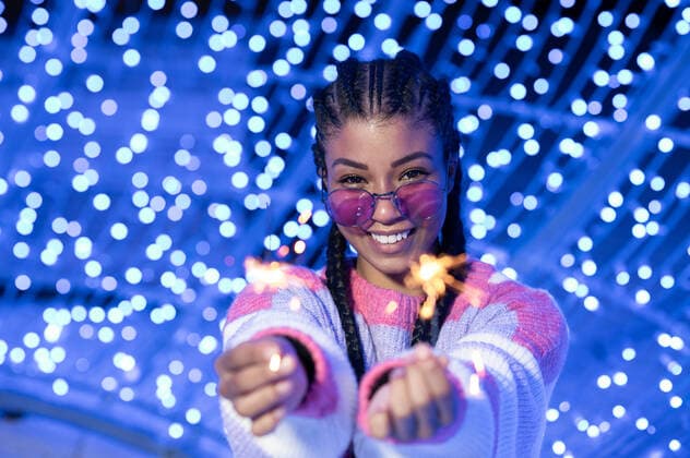 Happy young African American female in trendy sunglasses in warm sweater holding burning Bengal lights and looking at camera with smile while standing against blurred glowing garlands, Model released Copyright: xSergioxMenax MR