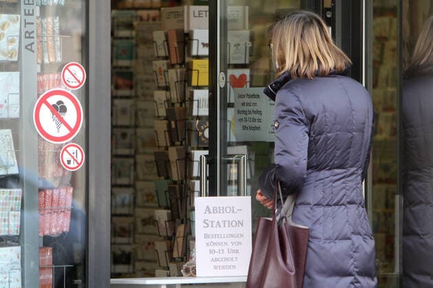 Schild mit Aufschrift Abholstation Bestellungen können von 10 - 13 Uhr abgeholt werden steht auf einem Tisch im Eingangsbereich eines geschlossenen Geschäfts in der Innenstadt von Hofheim, Hessen, Deutschland. Eine Kundin bei der Übergabe ihrer vorbestellten Ware vor der Tür.