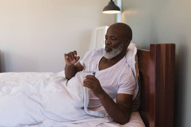 IMAGO Creative, Gesellschaft, Leisure Sickness Senior man taking medicine with a glass of water in bedroom Front view of senior African American man taking medicating with a glass of water in bedroom at home, model released, , property released, capetown, 26.02.2019, 1228659.jpg, senior citizen, senior, aged, ageing process, retired, retirement, pensioner, home, house, household, homey, apartment, domicile, abode, authentic, real life, bedroom, bed, blanket, nightwear, lying, holding, medicine, water, glass, treatment, ill, illness, sick, sickness, healthcare, healthcare and medicine, leisure, lifestyle 50s, Mature Adult, Man, Male, Black, 55-59 Years, Mature Men, African Ethnicity Indoors,Head and shoulders