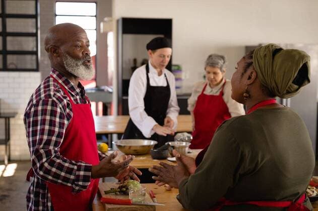 Chefs discussing while cooking Side view of a multi-ethnic group of Senior adults at a cookery class, an African American couple standing at a wooden table of ingredients talking in the foreground, and a Caucasian woman listening to instructions from a Caucasian female chef wearing chefs whites and a black hat and apron in the background, model released, , property released, Cape Town, South Africa, 05.12.2019, 1317530.jpg, people,discussing,discuss,gesturing,gesture,dough,rolling out,work station,team,together,chefs whites,uniform,counter top,real people,restaurant,business,food,kitchen,cookery,cuisine,culinary,catering,profession,job,experience,skill,busy,service,professional,confident,work,worker,working,cooking,preparation,preparing,ma