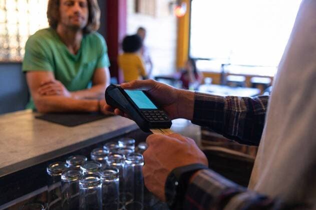 Man paying at the counter with his card Side view mid section of a mixed race male barman standing behind the bar in a pub, holding a credit card reader and inserting a credit card into it for payment, with a Caucasian male customer standing and leaning on the bar in the background, waiting for his credit card to be returned after making the payment, model released, , property released, Cape Town, South Africa, 15.11.2019, 1322565.jpg, people,barman,service,technology,paying,payment,business,small business,credit card reader,credit card,holding,waiting,leaning,real people,friends,friendship,socialising,freedom,free time,fun,hanging out,relaxing,gen z,generation z,pub,bar,leisure,entertainment,weekend,casual,trendy,hip,drinking,interacting