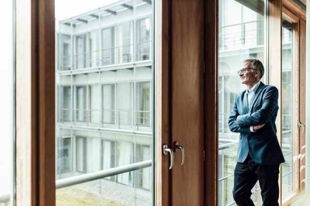 IMAGO Creative: Neue Aussichten Smiling male entrepreneur with arms crossed looking through glass in corridor model released Symbolfoto property released GUSF05323