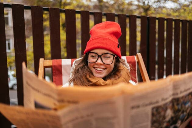 IMAGO Creative: Kulturelle Folgen der Pandemie: Gesundheitsbewußtsein und Alleinsein Smiling woman wearing knit hat and eyeglasses reading newspaper while sitting at sidewalk cafe model released Symbolfoto OYF00340