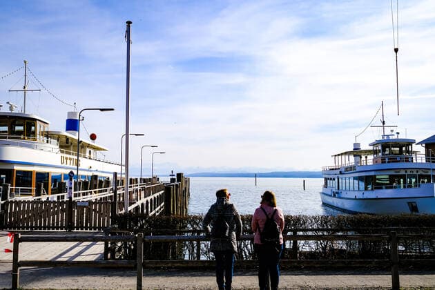 Frühlingswetter am Ammersee 25.03.2021: Ausflügler vor Schiffen bei Frühlingswetter im Hafen von Stegen (Inning am Ammersee; Oberbayern) mit Blick in Richtung Alpen. Inning Bayern Deutschland