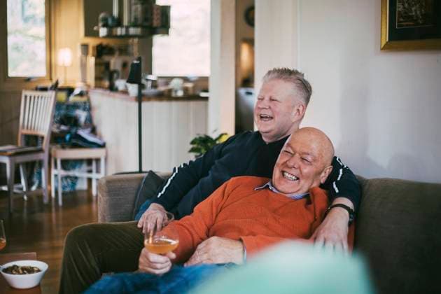 IMAGO Creative: Pride Month, Juni 2022, Pärchen und Familien Cheerful gay couple laughing while sitting on sofa in living room Sweden, Varmdo, model released, property released Copyright: xMaskotx , MA85505