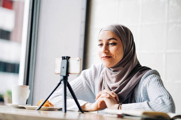 Low angle of cheerful Muslim female in headscarf shooting video on smartphone on tripod for blog while sitting at table in cafe Copyright: xJakexJakabx Model Released