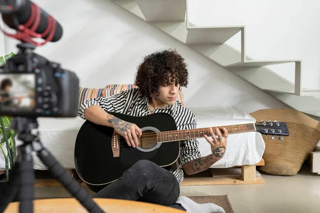 Young tattooed male guitarist playing acoustic guitar while recording video on photo camera in house room, Model released, Property released Copyright: xJosexCarlosxIchirox MR