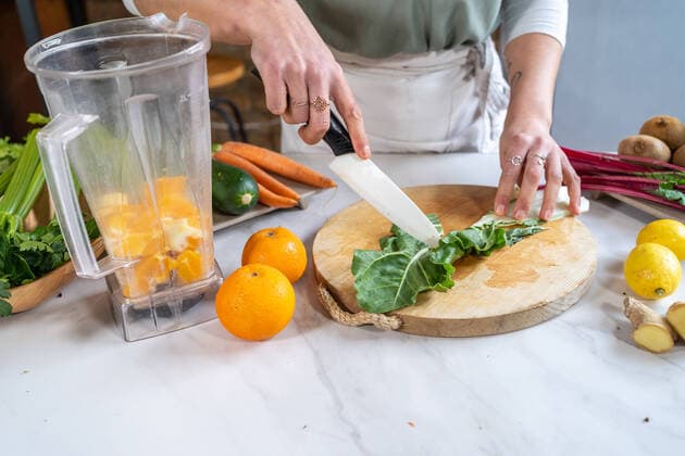 Cropped unrecognizable female cutting fresh chard leaves on chopping board against blender bowl with orange slices in house kitchen Copyright: xThexTrioxStudiox Model Released