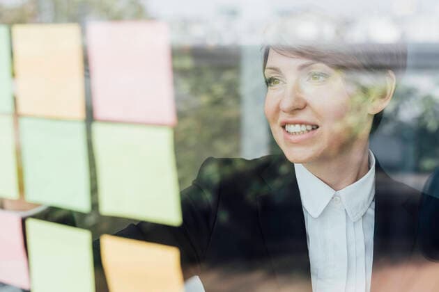 Smiling female entrepreneur looking at adhesive notes on glass wall model released Symbolfoto property released MEUF02264