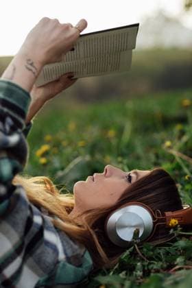 Side view of young attentive female in modern headset reading textbook while lying on meadow in summer, Model released Copyright: xEkaitzxGeriquex MR