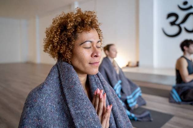 Side view of ethnic female wrapped in blanket meditating with Namaste hands in studio with diverse people practicing yoga together, Model released, Property released Copyright: xJordixBatallerx MR
