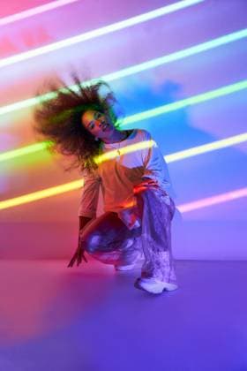 Full body stylish confident African American female dancer with curly hair and sunglasses crouched looking at camera in neon lights in dancing studio Copyright: xManuelxRuizx Model Released
